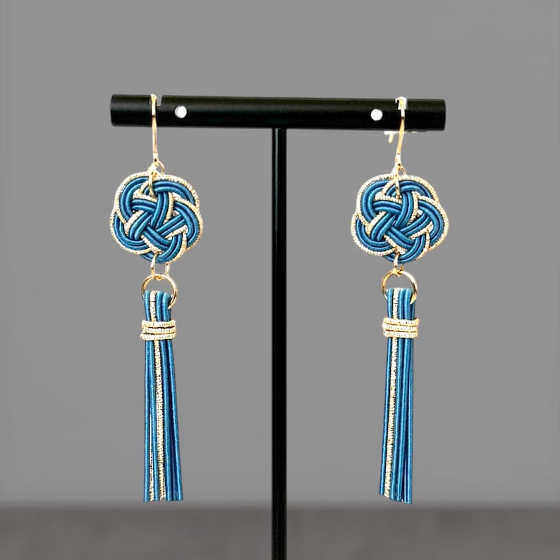 May include: A pair of blue and gold earrings with a tassel. The earrings feature a knotted design and are hanging from a black stand.
