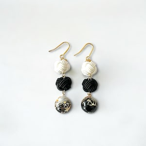 Può includere: Un paio di orecchini pendenti con tre perline ciascuno. La perlina superiore è bianca, la perlina centrale è nera e la perlina inferiore è una perlina con un motivo nero e oro.