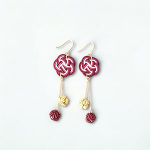 以下が含まれることがあります： ブルゴーニュ、白、ピンクの色合いの、花の結び目模様が特徴の手作りのイヤリングのペア。イヤリングには、金色のフックと、金とブルゴーニュ色の球体を含む、ぶら下がっているアクセントが付いています。イヤリングは白い背景に表示されます。