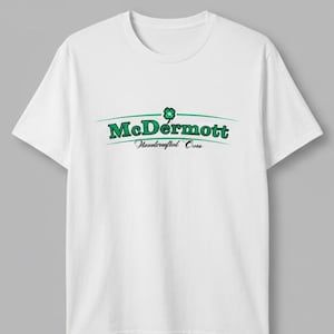 May include: A white short-sleeve t-shirt featuring a green graphic on the chest. The graphic displays the text McDermott in a bold green font, topped with a small green four-leaf clover icon, and the text Handcrafted Cues in black script below.