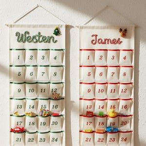 May include: Two hanging fabric advent calendars with twenty-four pockets each. The left calendar features the name Weston in green with a small tree icon. The right calendar features the name James in red with a reindeer icon. Both have numbered pockets.