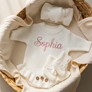 May include: A white baby outfit set with the name "Sophia" embroidered in pink. The set includes a sweater, headband, and mittens, arranged in a woven basket with a soft, cream-colored blanket.