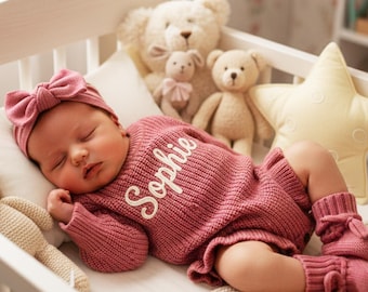 May include: A sleeping baby in a white crib wearing a pink knitted outfit with the name "Sophie" embroidered on the front. The outfit includes a matching headband, socks, and shorts. The crib is decorated with stuffed animals and a star-shaped pillow.
