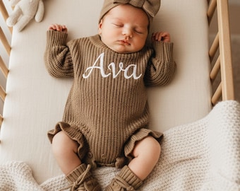 May include: A brown knitted long-sleeve romper with the name Ava embroidered in white script on the chest, paired with a matching brown bow headband and brown knitted booties. A small cream-coloured plush rabbit toy lies nearby.