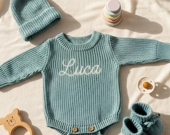 May include: A collection of baby items in a soft blue hue, including a knit romper with "Luca" embroidered on the front, a matching beanie with a pom-pom, booties, and a wooden bear teether. A pacifier and a stacking toy are also included.