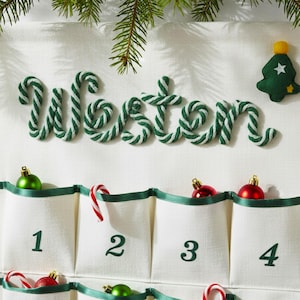 Pode incluir: Um calendário do advento de tecido branco com o nome Weston escrito em corda trançada verde e branca. O calendário possui bolsos numerados de um a quatro, decorados com pequenos enfeites verdes e vermelhos e bengalas doces.