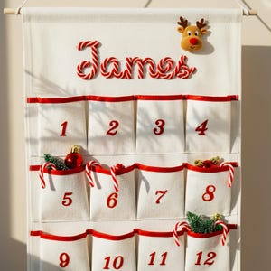 May include: A cream-colored fabric advent calendar hanging from a wooden rod, featuring the name James in red and white candy cane lettering, a plush reindeer head, and twelve pockets with red trim and red numbers containing small holiday ornaments.