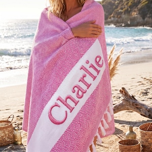 May include: Pink and white patterned beach towel with the name "Charlie" embroidered in pink. The towel has a diamond pattern and white fringe. The towel is wrapped around a person on a beach.