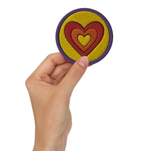 Può includere: Una toppa ricamata di colore viola e giallo con un motivo a forma di cuore rosso, arancione e giallo.
