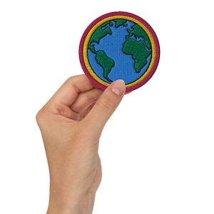 May include: A hand holding a round embroidered patch with a blue and green globe design. The globe is surrounded by a yellow and pink border.