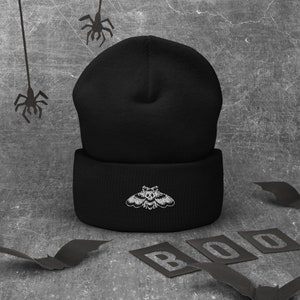 May include: Black beanie with a white embroidered moth design. The beanie is folded down and has a thick cuff.