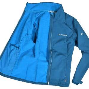 May include: A blue jacket with a full zip, showing a lighter blue interior. The jacket features a small logo on the chest and sleeve. It has two internal pockets. The jacket is made of a soft, flexible material.