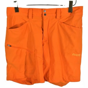 May include: Bright orange outdoor shorts with multiple pockets and a zipper closure. The shorts have a button and zipper closure at the waist and a small logo on the right leg. The shorts are hanging from a black hanger.