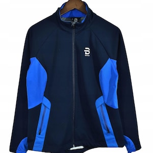 May include: Navy blue and bright blue athletic jacket with a full zipper closure. The jacket features a stand-up collar, two front pockets, and a white logo on the chest. The jacket is hanging on a wooden hanger.