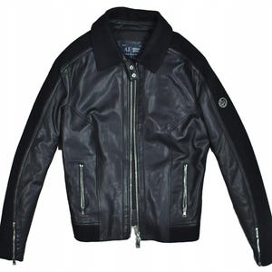 May include: Black leather jacket with a zip-up front, two side pockets, and a black collar. The jacket has black fabric panels on the sleeves and a logo on the upper arm. The jacket is a classic style.