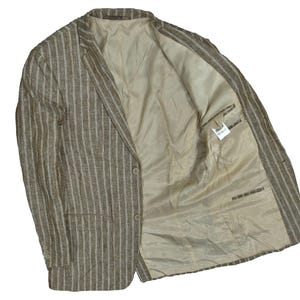 Puede incluir: Un blazer a rayas marrones y blancas con cierre de un solo botón. El blazer tiene un forro interior de color beige claro y una solapa con muescas. La chaqueta tiene dos bolsillos delanteros y tres botones en cada puño.