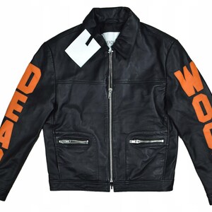 Puede incluir: Chaqueta de cuero negra con letras naranjas "DOOM WOOD" en las mangas. La chaqueta tiene cuello, cierre de cremallera y dos bolsillos delanteros con cremalleras. Una etiqueta blanca está adherida cerca del cuello.