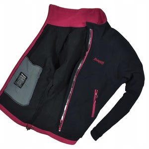 May include: Black and magenta Bergans jacket. The jacket has a magenta collar, zipper, and pocket accents. The interior is black, with a gray pocket on the left side. The Bergans logo is visible on the chest. The jacket is open, displaying the interior.