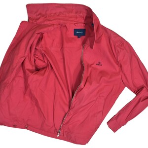 May include: A red jacket with a full zip, a stand-up collar, and long sleeves. The jacket features the brand name "GANT" on the chest and inside the collar.