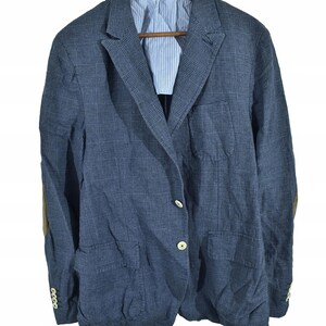 May include: A blue plaid blazer with elbow patches and cream-colored buttons. The blazer has two front pockets and a light blue and white striped lining visible at the collar. The jacket is hanging on a wooden hanger.