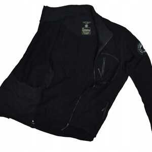 Puede incluir: Chaqueta negra con cremallera, cuello alto y múltiples bolsillos. La chaqueta presenta un parche con el logotipo en la manga y un pequeño logotipo en el pecho. La chaqueta está hecha de un material suave, posiblemente similar al forro polar.