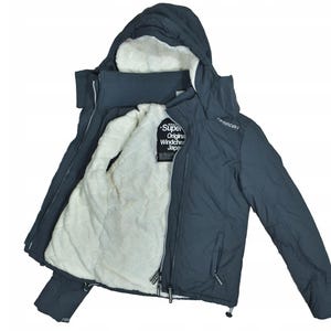 Peut inclure: Veste à capuche bleu foncé avec une doublure intérieure duveteuse de couleur crème. La veste a une fermeture éclair et une étiquette avec du texte. La veste est ouverte, révélant la doublure douce et un col côtelé.