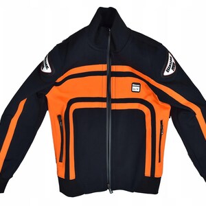 Può includere: Giacca con zip blu navy e arancione. La giacca presenta strisce e pannelli arancioni, un collo alto e una piccola toppa con logo. Le maniche hanno strisce arancioni e la giacca ha una cerniera nera e polsini neri.