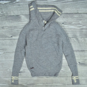 Puede incluir: Un suéter de punto gris con cuello con cremallera y textura acanalada. El cuello y los puños presentan rayas color crema. Una pequeña etiqueta está cosida en el lado inferior izquierdo. El suéter está sobre una superficie de madera.