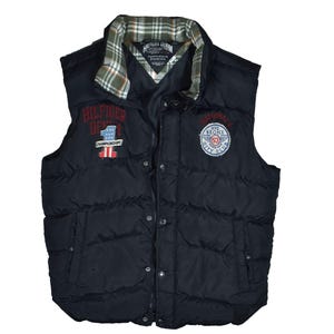 Peut inclure: Veste matelassée noire avec col à carreaux et plusieurs écussons. La veste présente le texte "HILFIGER DENIM" et "NATIONALS NYC BRONX CITY CLUB". La veste a une fermeture boutonnée et deux poches.