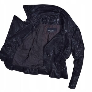 May include: A black leather jacket with a wide collar and button closure. The jacket is open, revealing a dark brown interior lining. The brand name, MARCCAIN, is visible on the inside. The jacket has a classic, tailored design.