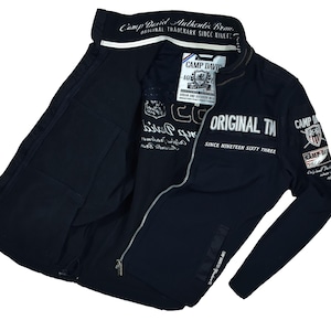 May include: A navy blue zip-up jacket with white lettering and patches. The jacket features the text "Camp David Authentic Brand" and "Original Trademark Since Nineteen Sixty Three". The jacket has a full zipper and two front pockets.