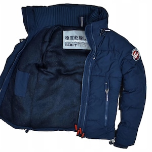 May include: Navy blue puffer jacket with a high collar and a zip-up front. The jacket features a quilted design, a small pocket, and a patch on the sleeve. The interior lining is black, and the jacket has the text "SUPER SOFT TECH".