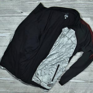 Puede incluir: Chaqueta deportiva negra con una capa interior reflectante plateada. La chaqueta tiene cremallera completa, mangas largas y detalles rojos en el hombro derecho. La chaqueta está sobre una superficie de madera gris claro.