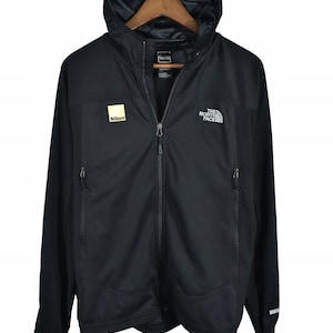 May include: Black hooded jacket with a full-length zipper, featuring the Nikon logo on the chest and "THE NORTH FACE" logo on the right chest. The jacket has two zippered pockets and a mesh-lined hood. The jacket is hanging on a wooden hanger.