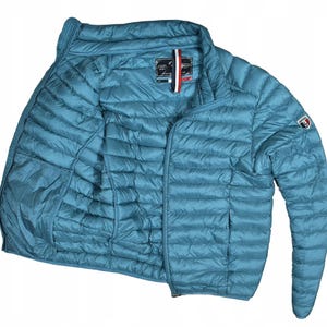 May include: Teal puffer jacket with a full zip. The jacket features a high collar and a small logo patch on the upper left sleeve. The quilted material and lightweight design make it suitable for cooler weather.