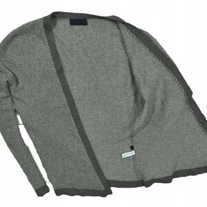 Op de afbeelding: Een grijze cardigan met een visgraatpatroon. De open trui heeft lange mouwen en een donkerdere grijze rand. De stof lijkt een zacht gebreid materiaal te zijn. De cardigan is open.