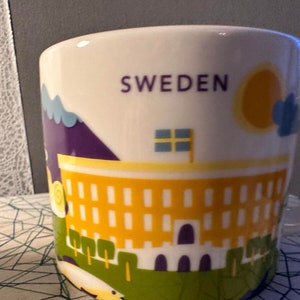 Może przedstawiać: Biały ceramiczny kubek z napisem "SWEDEN" i kolorowym wzorem. Wzór zawiera żółty budynek, słońce, chmury i flagę. Kubek ma zakrzywiony uchwyt.