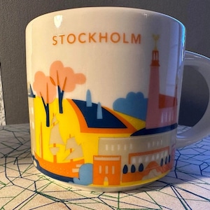 Może przedstawiać: Biały ceramiczny kubek z kolorową ilustracją Sztokholmu. Projekt zawiera budynki, drzewa i łodzie w odcieniach żółci, pomarańczy, błękitu i różu. Słowo "STOCKHOLM" jest wydrukowane na kubku w kolorze pomarańczowym.