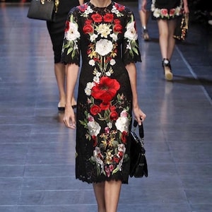 May include: A black knee-length dress featuring short sleeves and a high neckline, adorned with intricate, vibrant floral embroidery in shades of red, white, and green. The garment is crafted from lace fabric and displays a detailed botanical pattern.