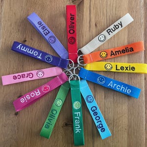 May include: A selection of fabric keyrings in various colours, each featuring a name and a smiling face. The keyrings are in shades like red, purple, pink, green, and blue, with white text displaying different names.