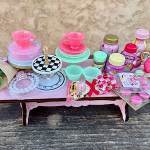 May include: A miniature pink table is set with a variety of dollhouse-sized items. There are pink teacups, plates, a teapot, and a cake. Other items include small jars, a bouquet of roses, and a clear container of macarons.
