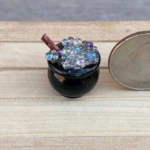 May include: A miniature black cauldron filled with iridescent blue and white beads, with a small wooden stick sticking out of the top.