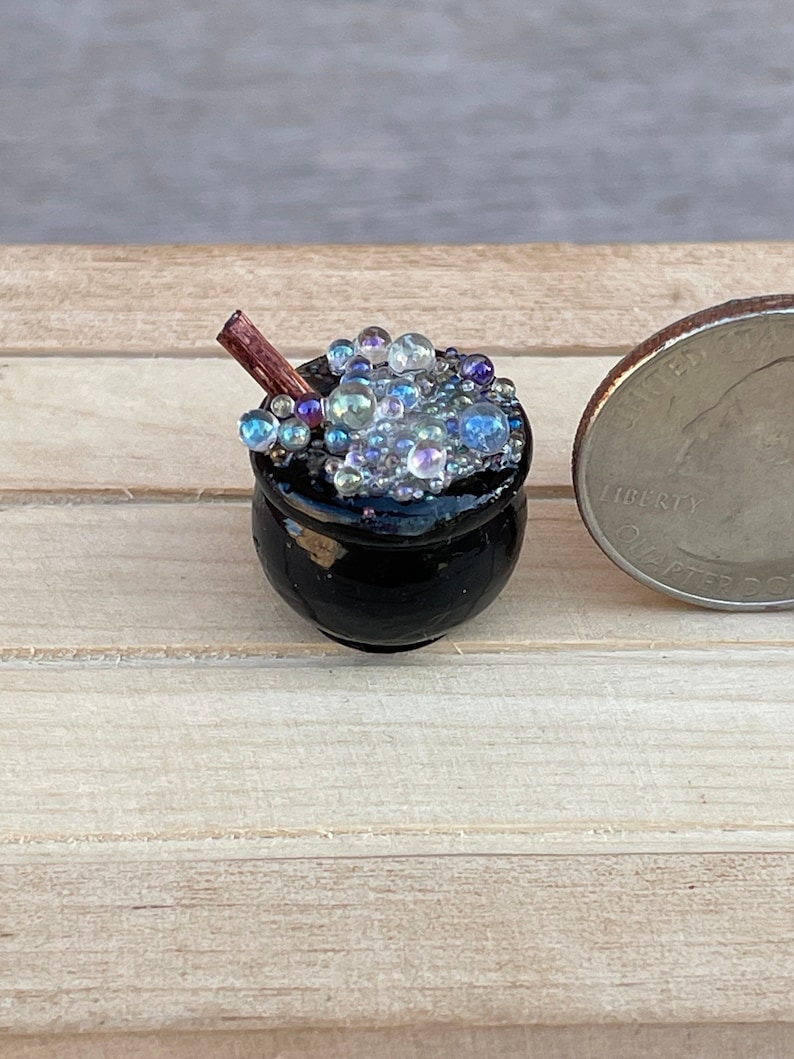 May include: A miniature black cauldron filled with iridescent blue and white beads, with a small wooden stick sticking out of the top.