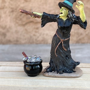May include: A black witch figurine with a green face and a black hat casting a spell with a wooden wand. The witch is standing next to a black cauldron with bubbling liquid.