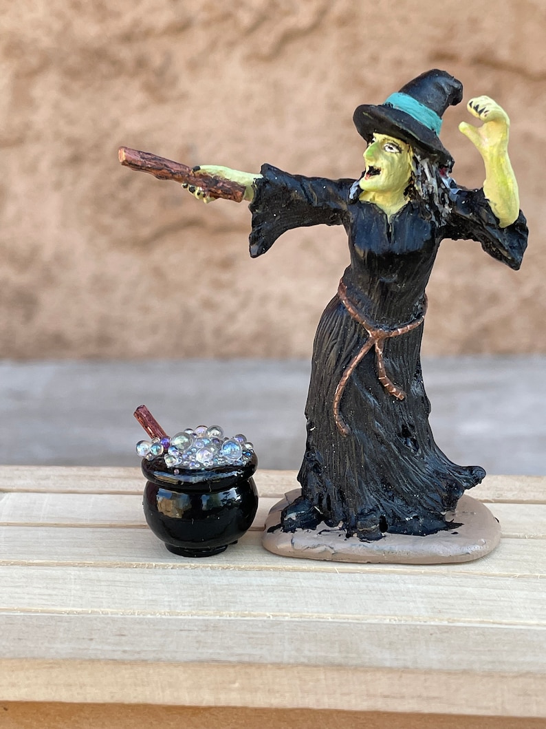 May include: A black witch figurine with a green face and a black hat casting a spell with a wooden wand. The witch is standing next to a black cauldron with bubbling liquid.