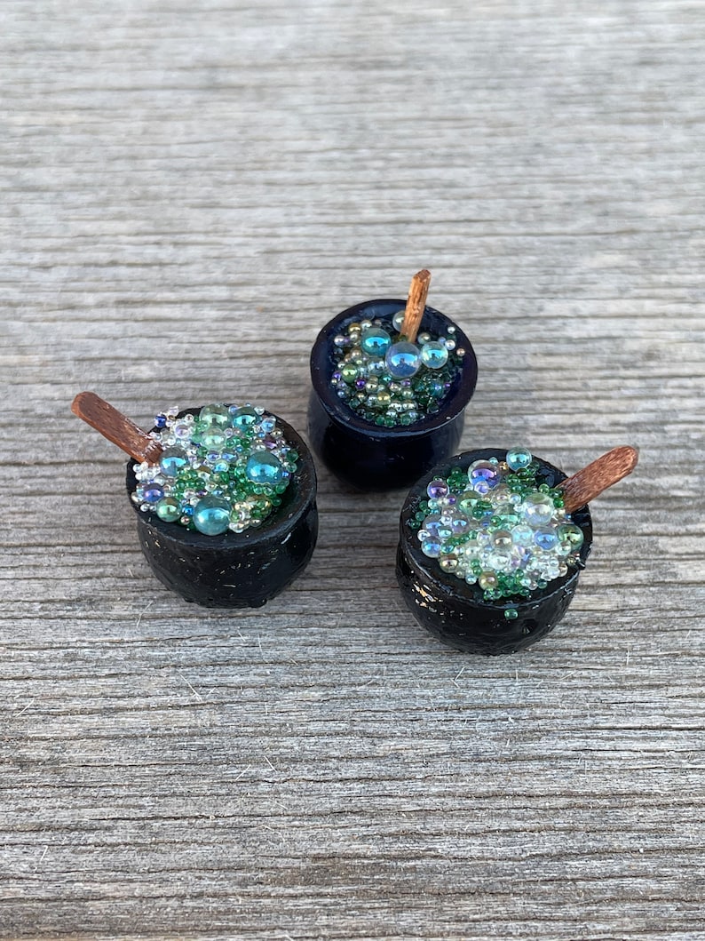 May include: Three miniature black cauldrons filled with green and blue iridescent beads, each with a small wooden stirring stick.