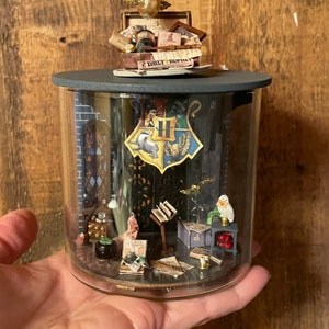 May include: A glass jar with a black lid containing a miniature Hogwarts scene. The scene includes a Hogwarts crest, a book, a cauldron, and a small owl figurine. The jar is sitting on a hand.