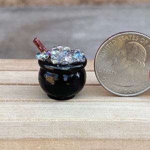 May include: A miniature black cauldron filled with iridescent bubbles and a small wooden stirring stick. The cauldron is sitting on a wooden surface next to a US quarter for scale.