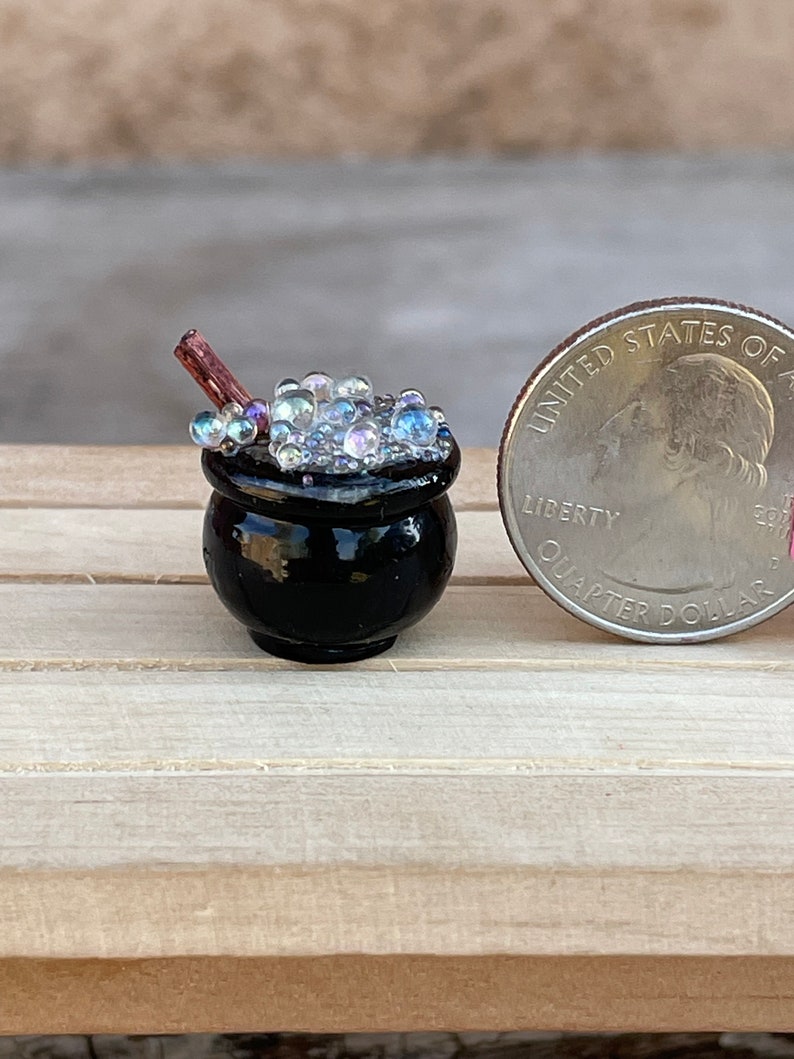 May include: A miniature black cauldron filled with iridescent bubbles and a small wooden stirring stick. The cauldron is sitting on a wooden surface next to a US quarter for scale.
