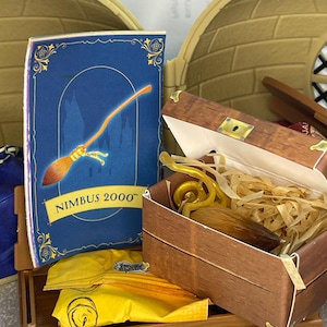 May include: A miniature toy set featuring a blue book with "Nimbus 2000" text and a golden broom illustration. A wooden chest contains a golden snitch and other accessories. A yellow pouch and other items are also visible.